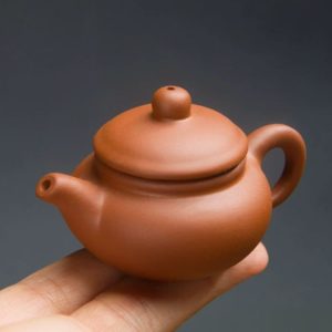 Миниатюрный чайник из пурпурной глины (чайный питомец), расположенный на указательном и среднем пальцах руки для демонстрации размера.